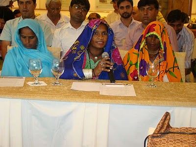 Hindi women sitting at a table. One of the women is talking into a microphone.