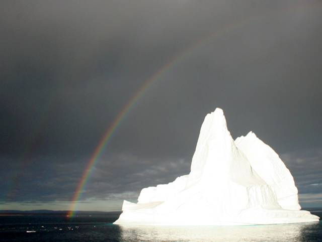 Cloudy skies and double rainbows over a bright white iceberg in Scoresby Sound, NE Greenland National Park.
