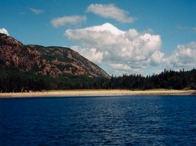 Beach at Acadia with water in foreground, mountains and cloudy skies in background