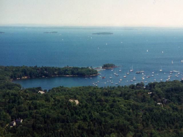 Ocean with inlet and forested land in foreground, small islands in the distance.  Sailboats anchored in the inlet and sailing in the distance