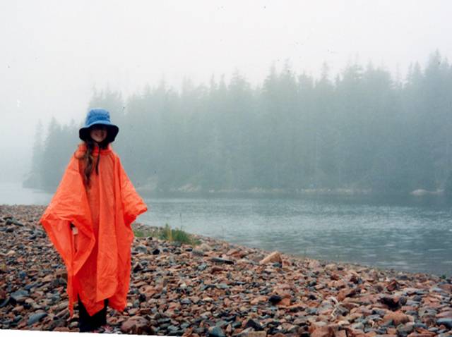 Person standing in the rain on rocky beach, wearing orange rain poncho and blue rain. Water and Evergreens in the background