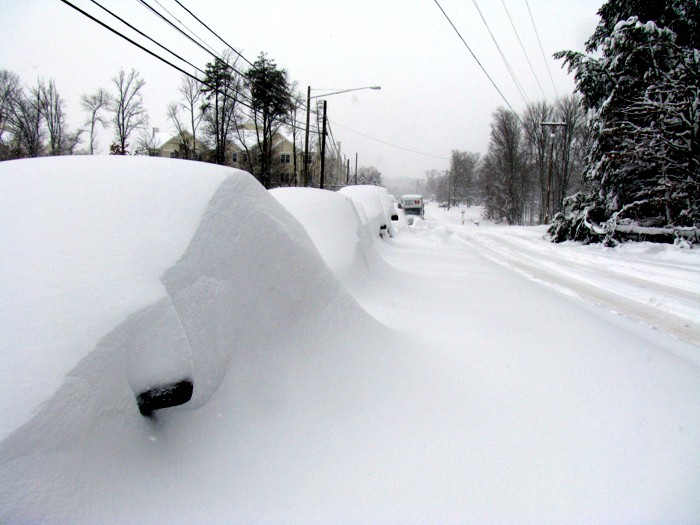 A photo of a snow-covered road with snow-covered cars parked alongside. 