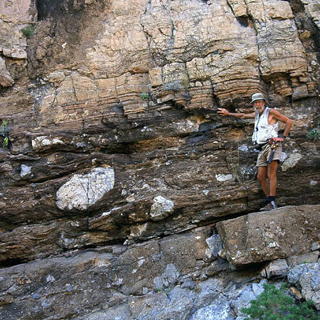 man standing against rock formation with horizontal rock layers. The top half is light tan the bottom half is brown grey