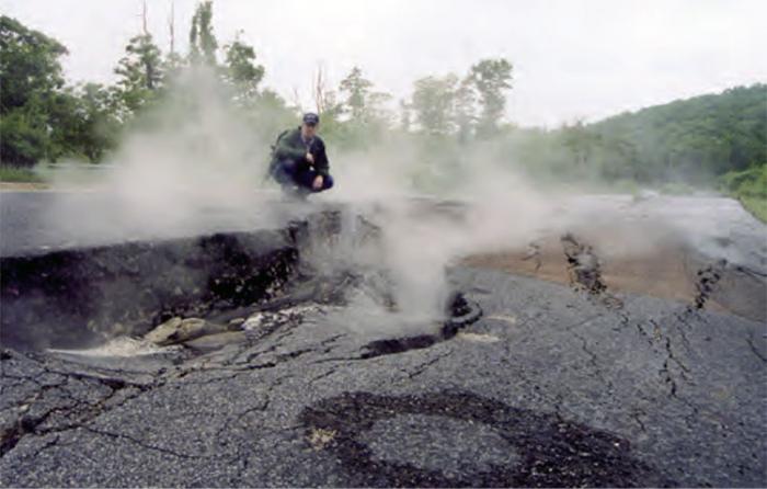 Person sitting on asphalt with big cracks in it watching the gases from an underground coal fire rising from the Earth’s surface.