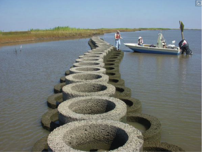Concrete cylinders placed along shoreline, and a person standing on cylinder near a small fishing boat.