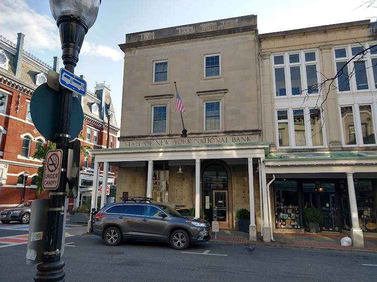 The image shows an urban intersection. The focus of the image is a building standing on the corner: a boxy three-story edifice faced in light colored stone. A metal canopy covers the first story as part of a portico that lines the street. Just above the canopy is an inscription that reads “STATE OF NEW YORK NATIONAL BANK,” and just above that, an American flag hangs from a pole.