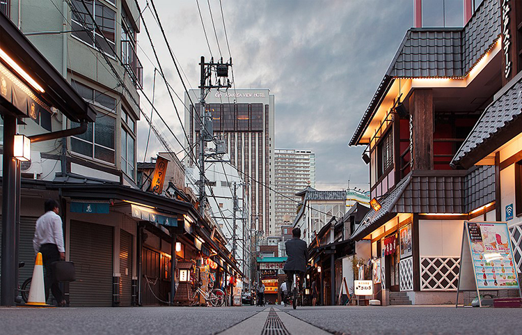 The image depicts a street, taken from ground level. On either side of the street are low (one- and two-storey buildings) featuring shops and restaurants. A man rides a bicycle down the street, away from the viewer, and a second person riding a bicycle is visible down the street. In the distance stand two skyscrapers against the cloudy sky. Architectural elements and signage suggest this is in Japan.