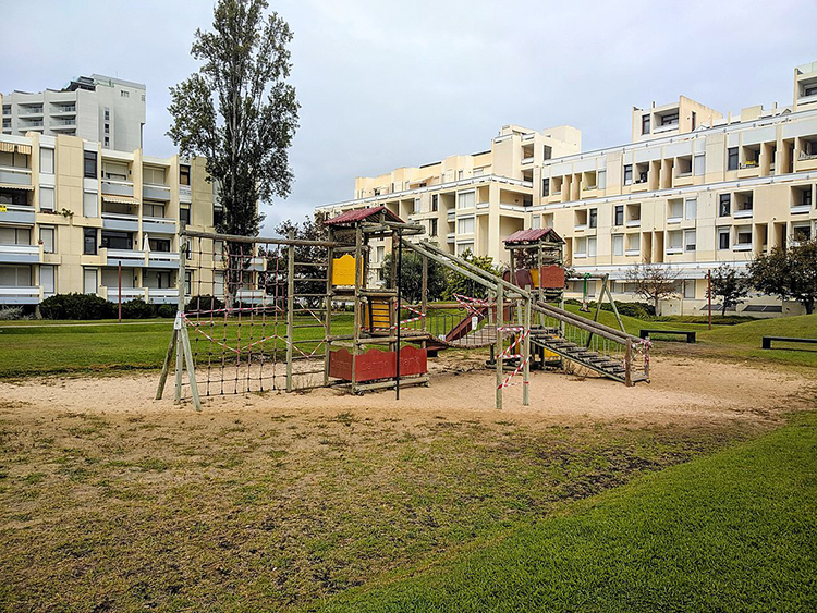 The image shows an empty playground dominated by a large, wooden, multi-use structure featuring ladders, a rope net, stairs, and towers. In the background stand two broad apartment buildings of four and five stories, and a third apartment tower in the distance.