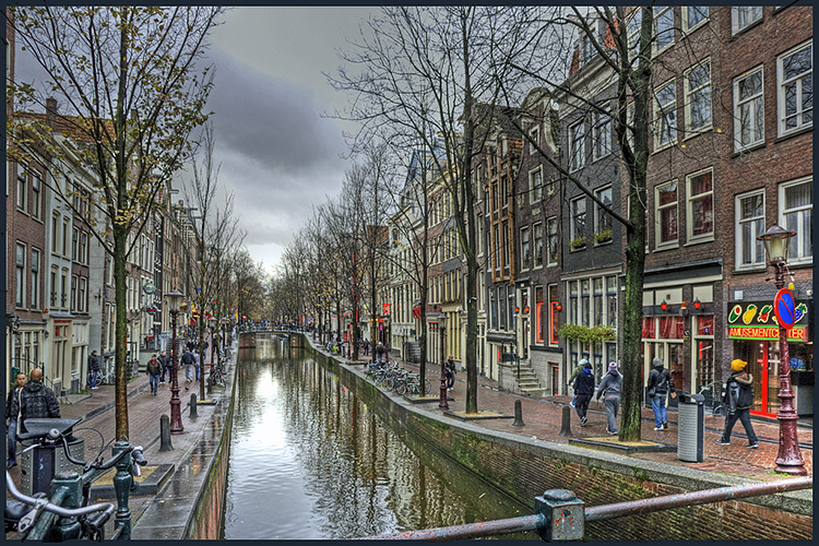 The image shows a brick-paved pedestrian street split by a canal down the center. On either side of the street are three-story row houses. Some of the facades feature red lights on sconces, and some windows are lined in red neon lights. Bicycles are parked along parts of the canal, and about two dozen figures (who all appear male) are visible walking along the street. In the distance, a pedestrian bridge crosses the canal. In the lower right-hand corner, a sign reading “AMUSEMENTCENTER” hangs on one facade.