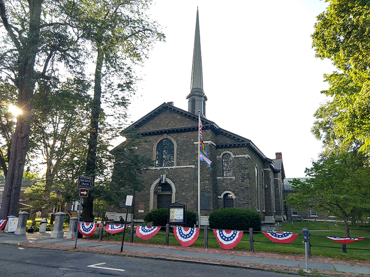 The image shows a Renaissance Revival-style stone church. Headstones are visible in the small lawn around the building, and a flagpole stands out front, with several flags hanging from it. A low iron fence marks the boundary between the yard and the stone sidewalks; red-white-and-blue bunting hangs along the railings.