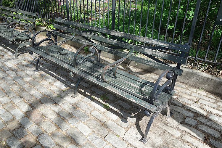 The image shows two park benches lining a stone walkway. The benches are long and consist of wooden slats with decorative iron feet and rails. Three rails divide the benches into five sections, making them too short for a person to lie down.