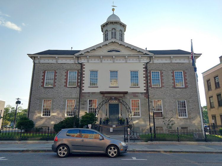 The image shows a Georgian style building in gray stone with a domed cupola. The building and its small yard are set off from the sidewalk by a low, wrought iron fence. At the near right corner of the building, a flagpole flies the flags of the United States and New York State. A car sits parked on the street in front of the building.