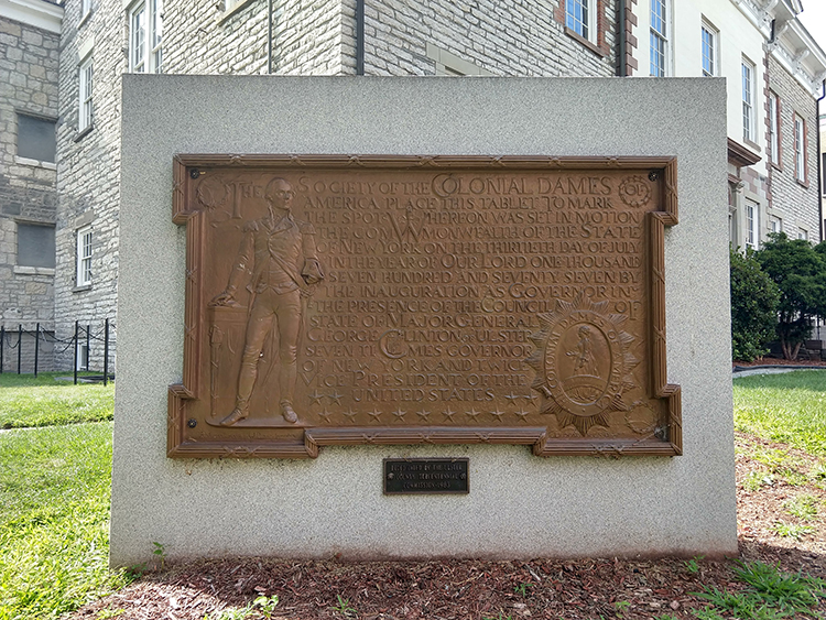 The image shows a standing stone monument bearing a large metal plaque. On the left side of the plaque we can see the standing figure of George Washington. In the lower right hand corner is the emblem of the Society of the Colonial Dames of America.