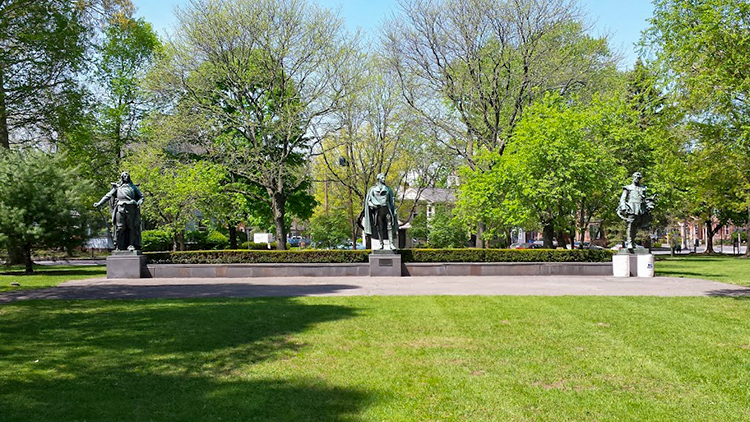 The image shows a section of a park, partly shaded by trees. A small plaza stands on a green lawn, bounded by a low wall on the far edge. Along the wall are three evenly-spaced plinths, upon each of which stands a bronze statue of a historic figure. Trees and the streetscape of Clinton Avenue are visible behind the statues.