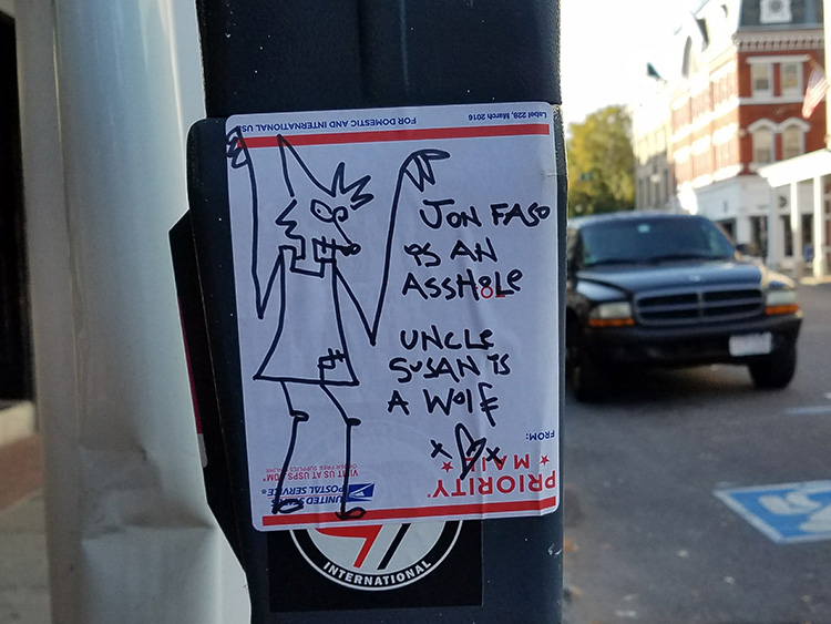The image shows a US Postal Service Priority Mail sticker on which someone has drawn a cartoonish figure of a wolf wearing a skirt. To the right of the drawing are several lines of handwritten text which read, “Jon [sic] Faso is an asshole” and “Uncle Susan is a wolf.” The sticker has been affixed to the side of a parking meter.