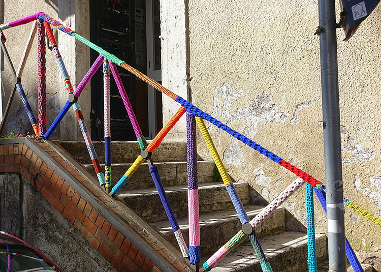 The image shows a set of exterior stairs. The iron railings of the staircase have been individually wrapped in a variety of colors of yarn. There are sections of blue, yellow, red, orange, pink, and green, among others.