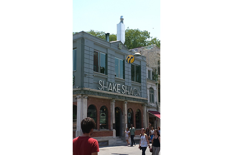 The image is a narrow shot showing a two-story building with the Shake Shack logo on it. In the foreground, a few people (men and women) are visible walking along the street.