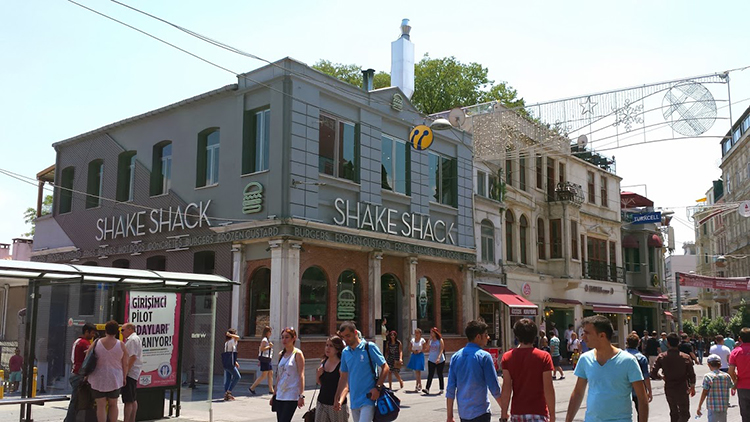 This image shows a broader shot of the Shake Shack in image 6.7a. A few dozen people walk down the street or stand in clusters, getting their bearings. The people are generally well dressed and clean cut. Other buildings, three or four stories tall, are visible, with facades that appear older than the one on Shake Shack. A sign in the upper right-hand corner reads TURKCELL while one in the lower left-hand corner reads “GİRİŞİMCİ PİLOT ADAYLARI ARANIYOR.”