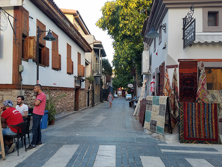 The image, taken from an intersection, shows a narrow street paved with brick and stone, running between two buildings. The buildings, no more than two or three stories in height, have facades of stone (lowest level) and plaster (upper levels), and functioning wooden shutters on the windows. In the left foreground, two young men sit at a table on the street while a third one stands with the group. In the right foreground is the corner of a shop that sells rugs, some of which are displayed on metal stands in front of the building. Small electric lamps with conical black shades are visible on either side of the street, situated at about the second floor of the buildings.