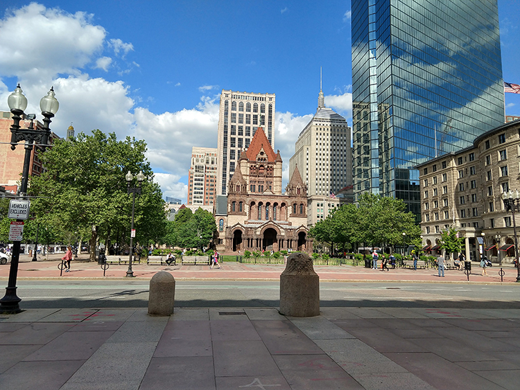 The image shows a plaza, at the far end of which is a church built in the Richardsonian Romanesque style. To the right of the church is a tall, modernist, glass skyscraper, and behind that is a square tower with a weather beacon on the top.