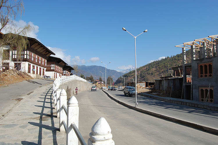 The image shows houses standing alongside a road, separated from it by a fence and retaining wall. The facades of the houses are whitewashed and adorned with multicolored wood framing the windows and upper register of the houses. The roofs have large, angled overhangs. Y-shaped street lamps and power lines run alongside the road. Mountains are visible in the distance.