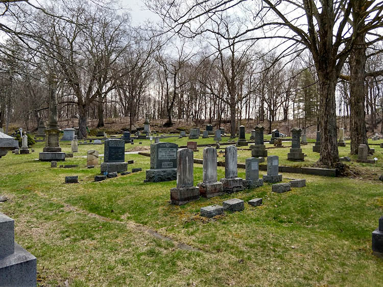 The image shows a grassy rural cemetery with clusters of headstones, obelisks, and other grave markers. Some are set off by stone curbs. The stones generally face the same direction.