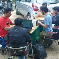 3 men sitting at a table with computers. The table is outside.