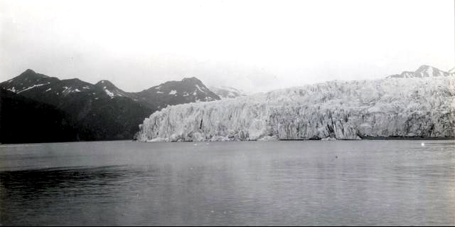 McCarty Glacier, AK, 1909. Large glacier in center, water in foreground, mountain visible to left in background.