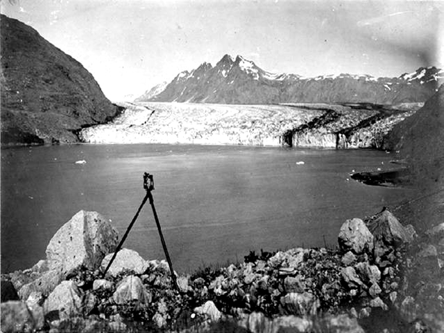 McCarty Glacier, AK, 1906. Rocky water’s edge and water in foreground. Glacier in background, in front of snow capped mountains.