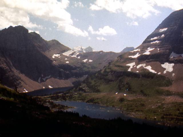 Glacier-carved scenery, Logan Pass, Glacier National Park. Snow-spotted mountains under cloudy skies, water in foreground.