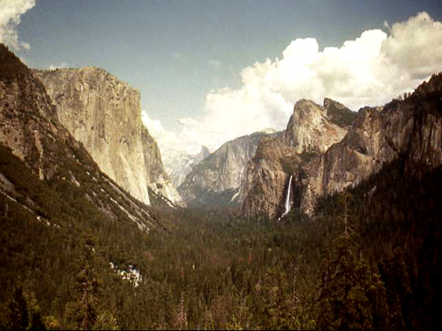 Yosemite valley under cloudy skies. Forest in the foreground and mountains in the background. Bridalveil Falls on right in distance.