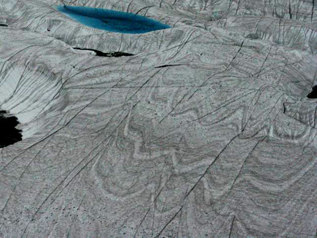 Ice sheet, NE Greenland National Park.  Wave-shaped folds are visible, showing flow of ice.  Bright blue meltwater lake visible, top left.