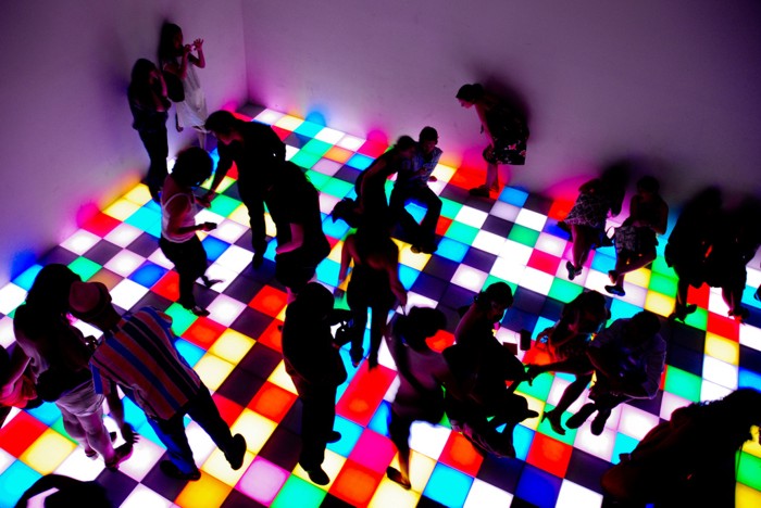 Photograph of a dance floor with kids standing, dancing, sitting and leaning against the walls. 