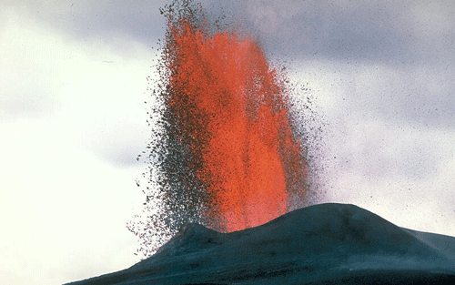 Tall fountain of lava, with black pieces coming off of the lava fountain. 